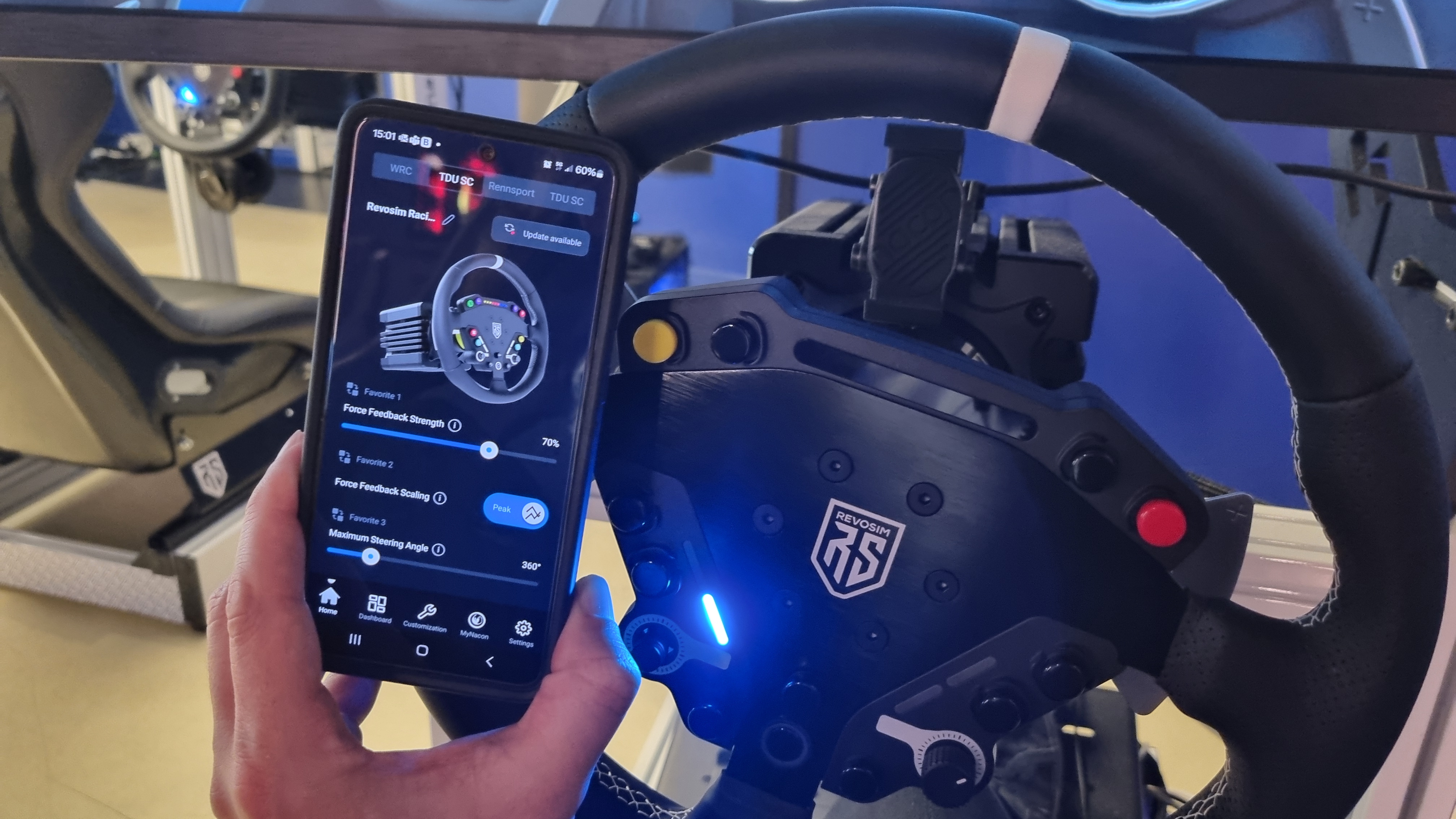 A phone being held next to a Nacon Revosim RS Pure racing wheel with an app on display showing adjustable settings.