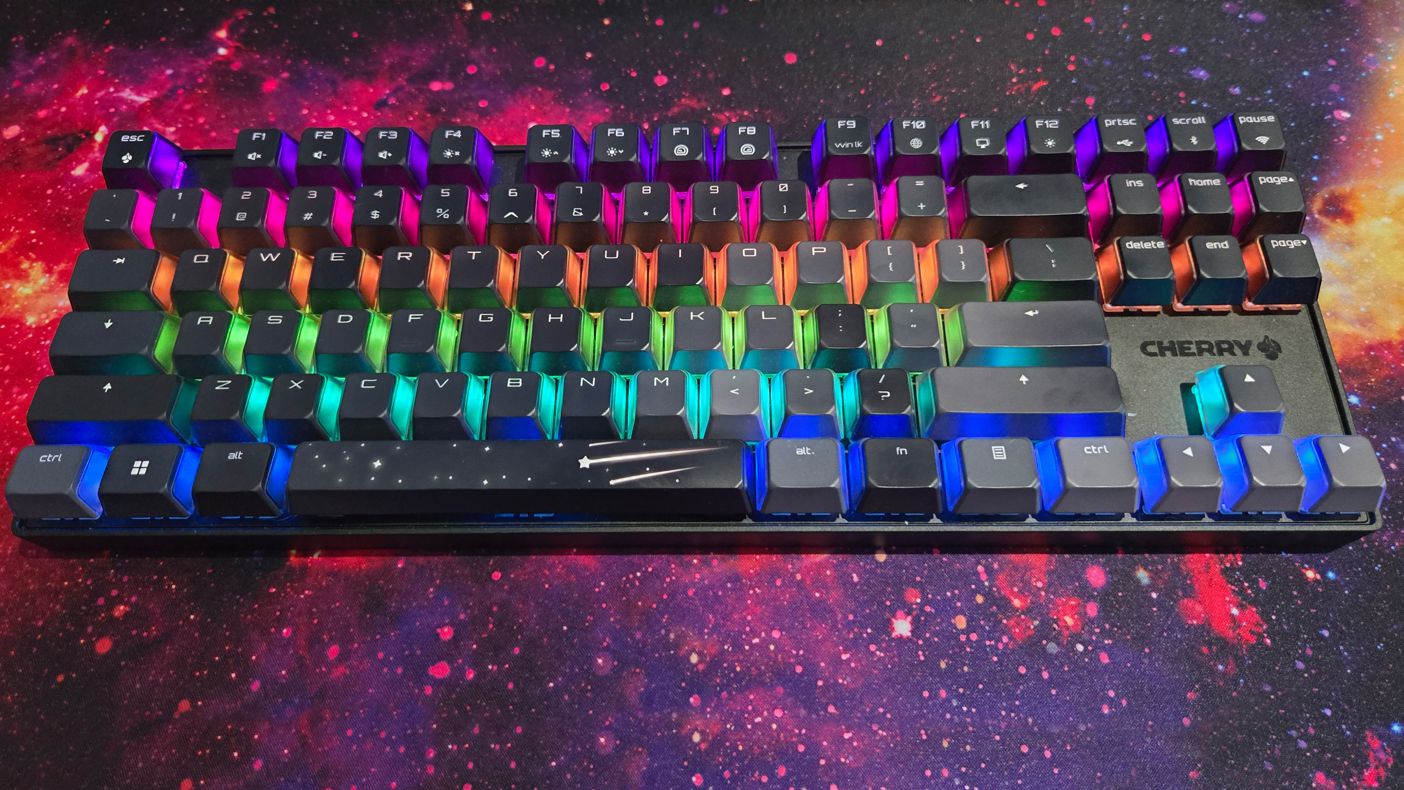 The Cherry MX 8.2 Wireless Xaga gaming keyboard sits on a large mouse mat depicting a nebula. This visual motif ties into the shooting star design on the keyboard's space bar. The keyboard's RGB lights are on, but the design on the space bar is opaque, so the RGB lights only shine through the frosted sides of the keyboard's alphanumeric keycaps.