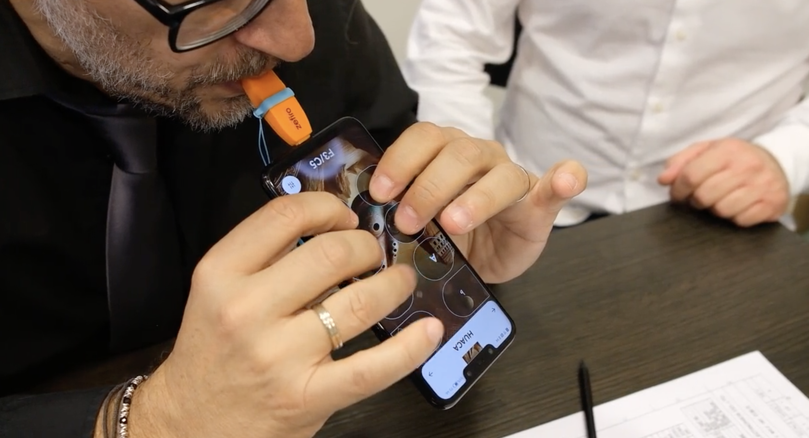 hap9jux2rzvzw8e7jme8cg-1 A man wearing glasses plays a Zefiro, attached to the bottom USB-C port of a smart phone. The phone offers touch screen buttons to play different notes.