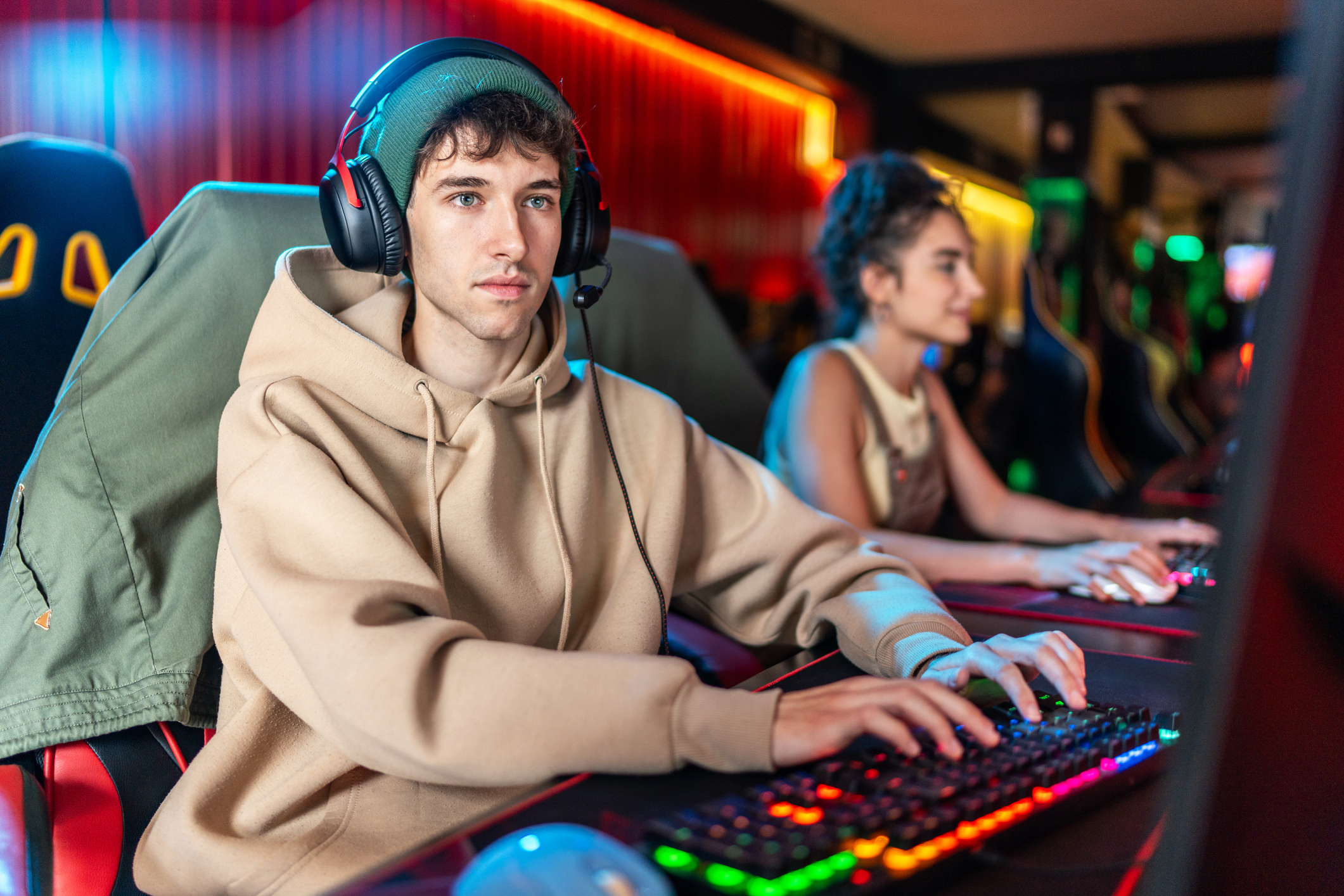 Young man concentrating on playing a video game, using a colorful keyboard and wearing headphones in an internet cafe, with another player visible in the background
